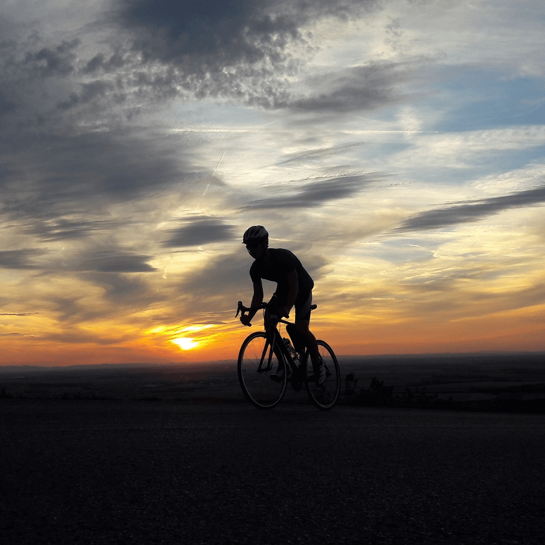 A cyclist is silhouetted against the fading light of dusk, preparing for a night ride on an open road. The darkening sky, with streaks of clouds, creates a serene yet dramatic atmosphere, perfect for evening or night cycling.